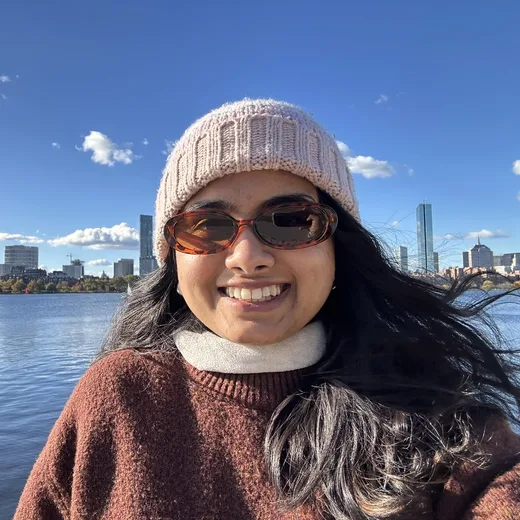 Rajmi smiling in a knit hat and sunglasses by the water with a city skyline behind